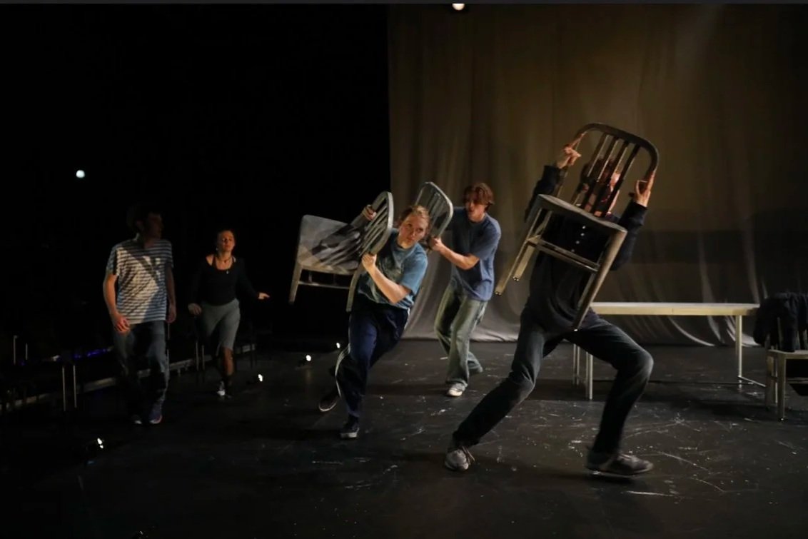 People practicing a dance or theater scene with chairs on a stage, some are holding chairs in dynamic poses.