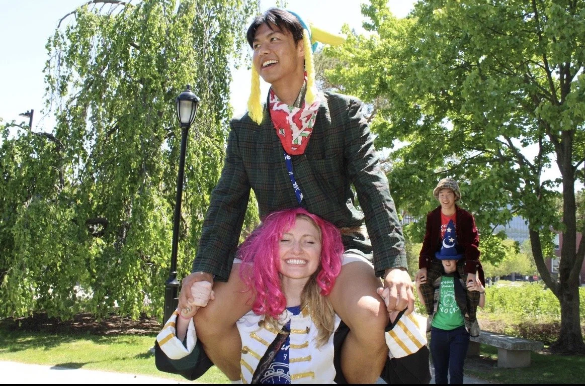 A group of people enjoying a sunny day in a park, with two individuals performing a piggyback ride and two others walking behind them. The scene is lively and cheerful with greenery and trees in the background.