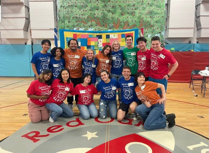 Group of young people posing inside a gymnasium, some kneeling in front and others standing behind, with a colorful quilt hanging on the wall behind them. They are wearing matching t-shirts in blue, red, and orange, each with a logo and the words "Graphics Tale Presents Clocks". The gym has a red and gray