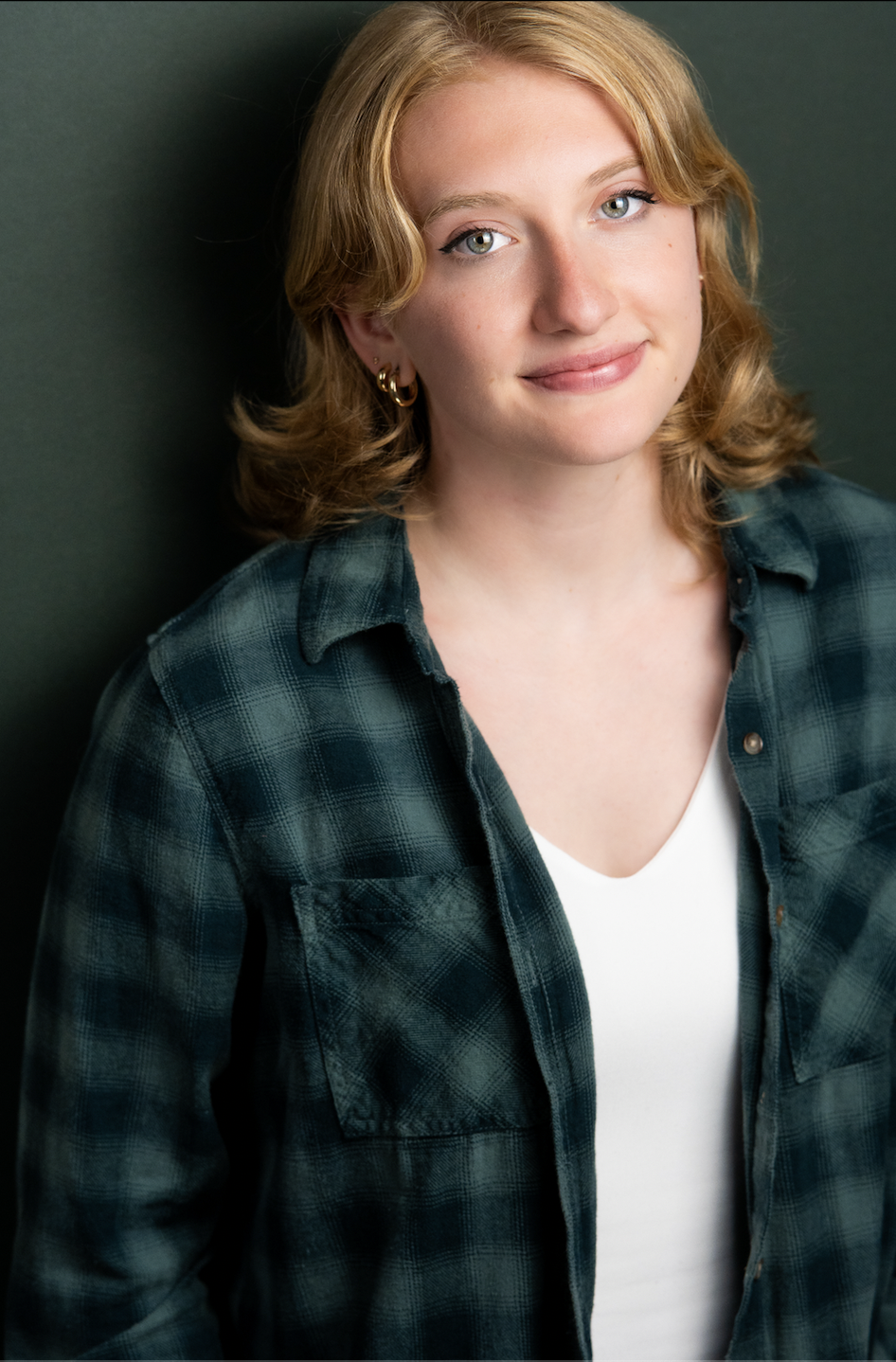 A young woman with shoulder-length wavy blonde hair, blue eyes, and earrings, wearing a dark green plaid shirt and white t-shirt, standing against a dark background.