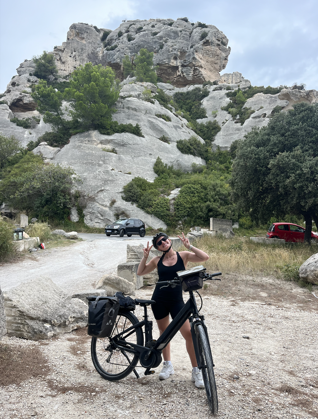 A woman in black athletic clothing and sunglasses standing next to a black bicycle, making peace signs with her hands, in a rocky outdoor area with a large rocky hill or mountain, trees, and a few parked cars in the background.