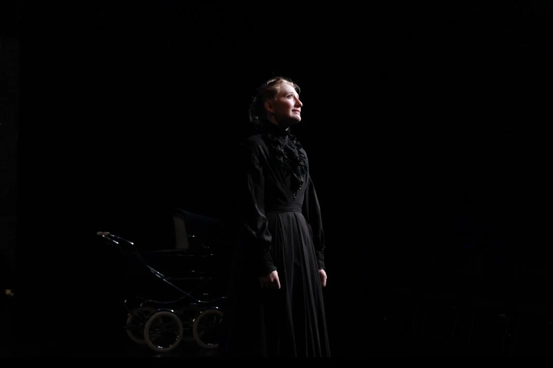 A woman in a black dress standing on stage with a spotlight on her, a wagon or stroller visible in the background.