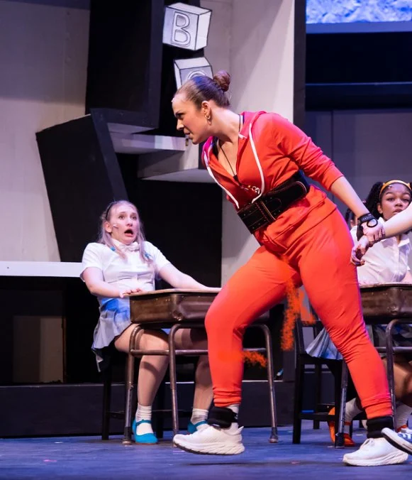 Actress in red tracksuit holding girls' hands during a school play scene on stage.