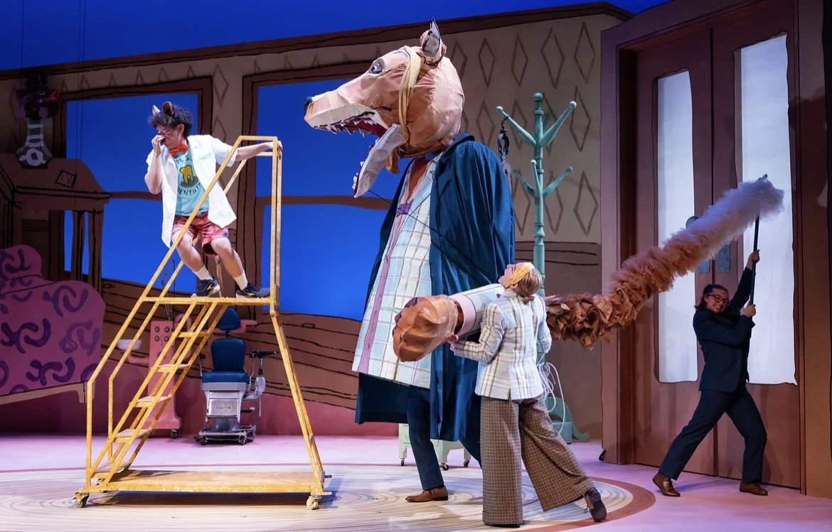 The image shows a theatrical stage scene with actors in animal costumes and colorful set design. One actor dressed as a mouse is standing on a yellow ladder, another actor with a horse head mask is in the center, and a third actor with a fox mask is holding a prop. A fourth person in a suit is pushing a large door with a fluffy smoke puff coming out.