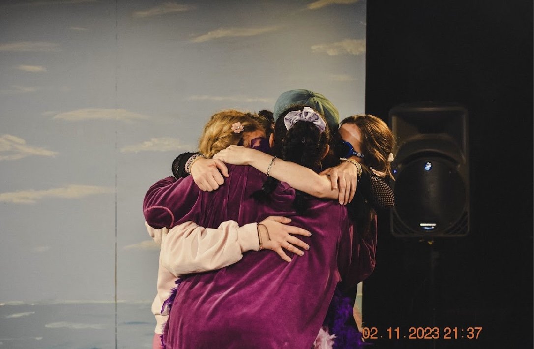 Group of four people hugging tightly on stage, with one person wearing a cap and others with long hair, against a painted sky backdrop, with a visible black speaker on the right.