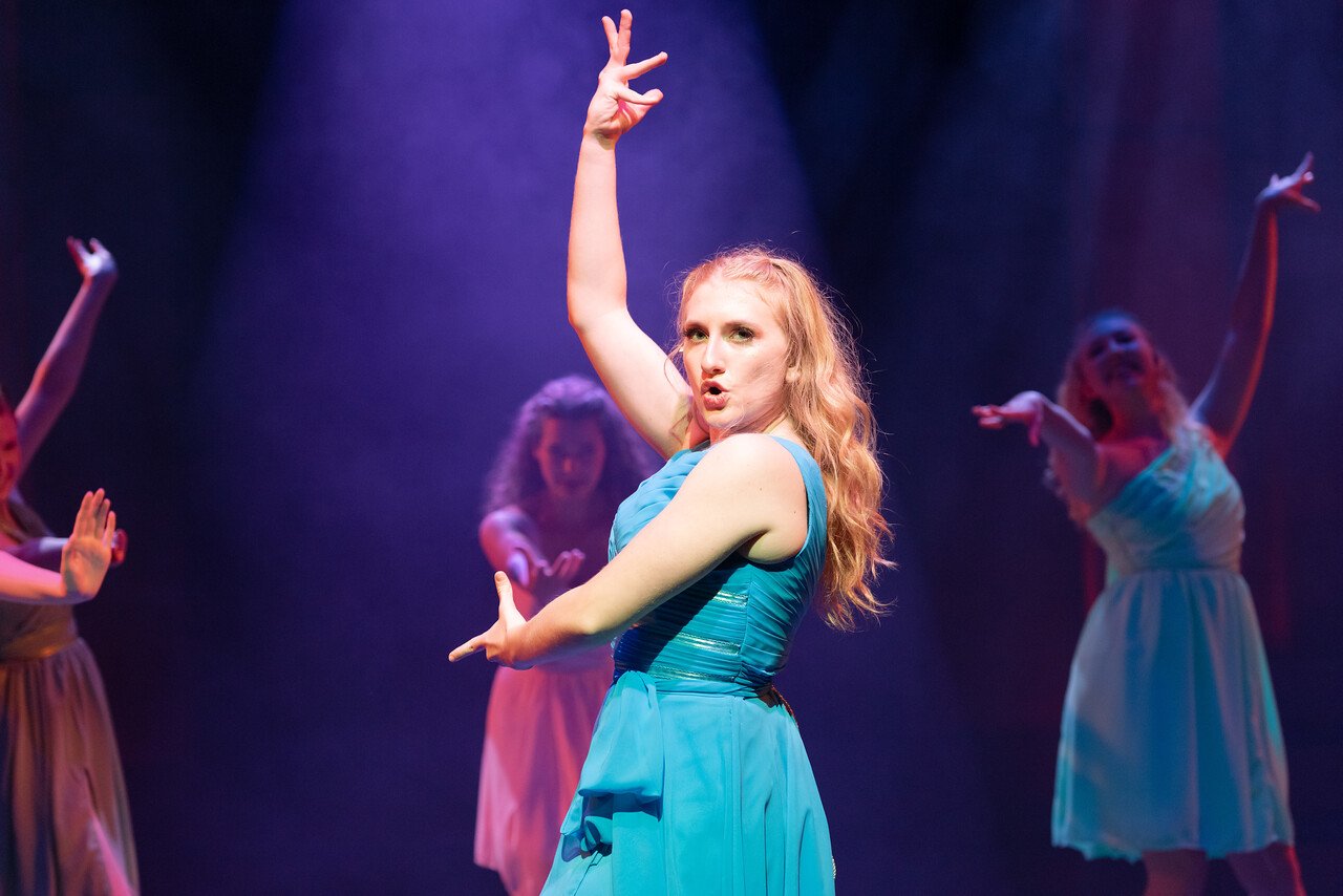 A woman with red hair in a blue dress performs a dance on stage, with three other women in similar dresses dancing behind her under purple stage lighting.