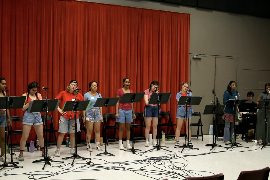 A group of young women singing on stage with music stands, microphones, and a red curtain backdrop.