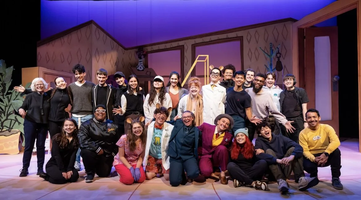 A group of actors on a stage, smiling and posing after a performance, with a set depicting a house interior in the background.