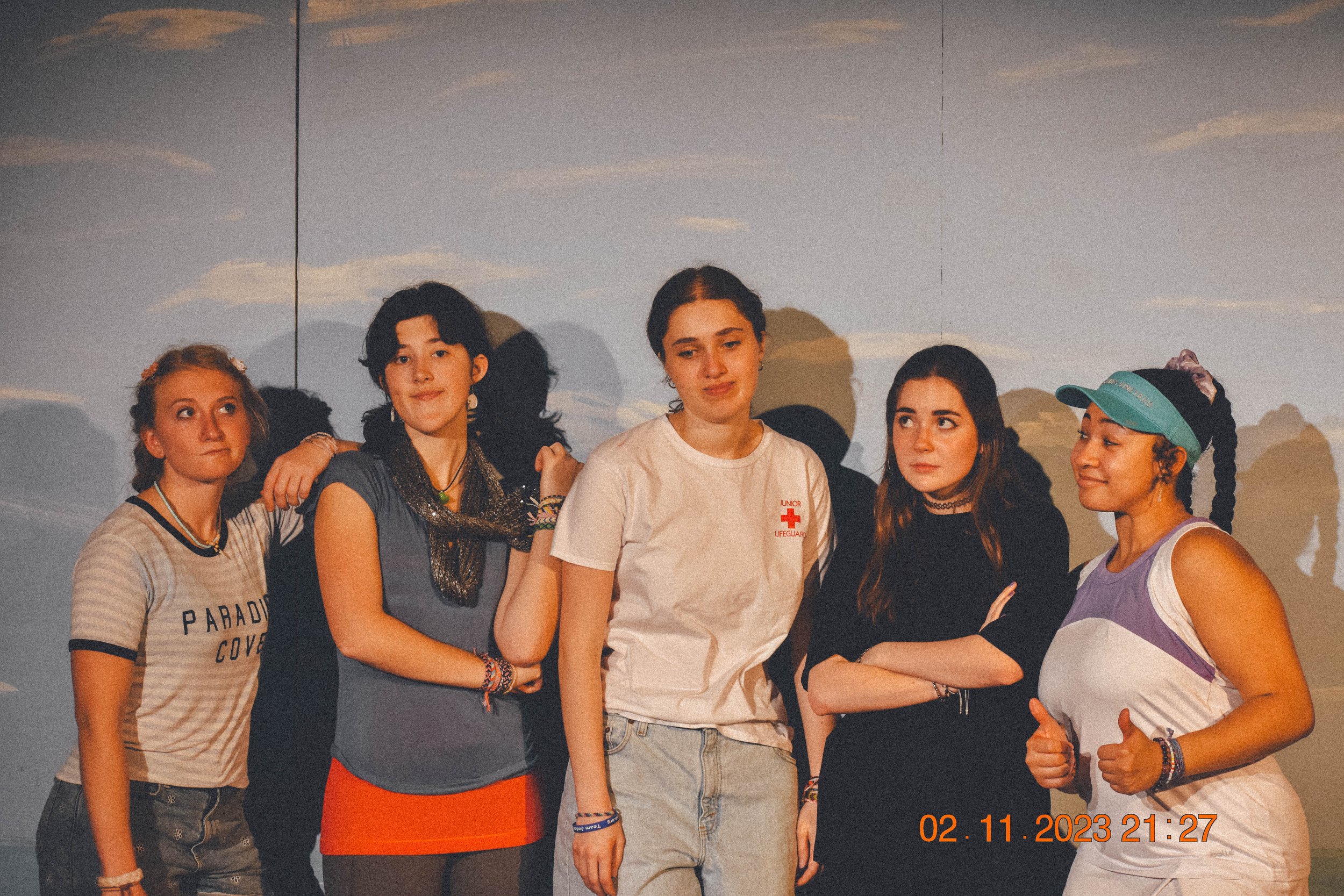 Five young women standing together against a backdrop that resembles a sky with clouds. They are dressed casually, with one in a white t-shirt with Red Cross emblem and others in various colorful outfits. They appear to be posing or gathered for a photo, with some smiling and some with serious expressions. The bottom right corner shows a timestamp, "02.11.2023 21:27."