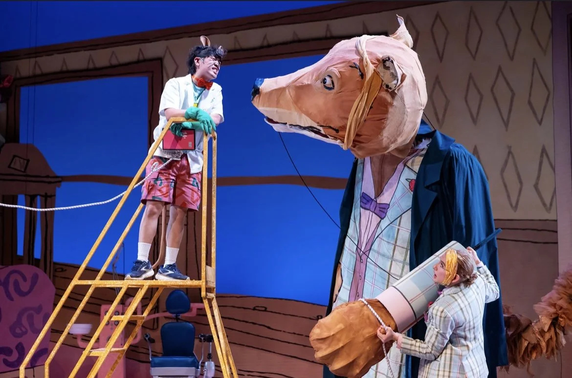 A theatrical performance featuring a person in a mouse costume with a large mouse head, a person with a painted face holding a cylindrical object, and a young boy on a ladder wearing props, all on a stage with painted set pieces.