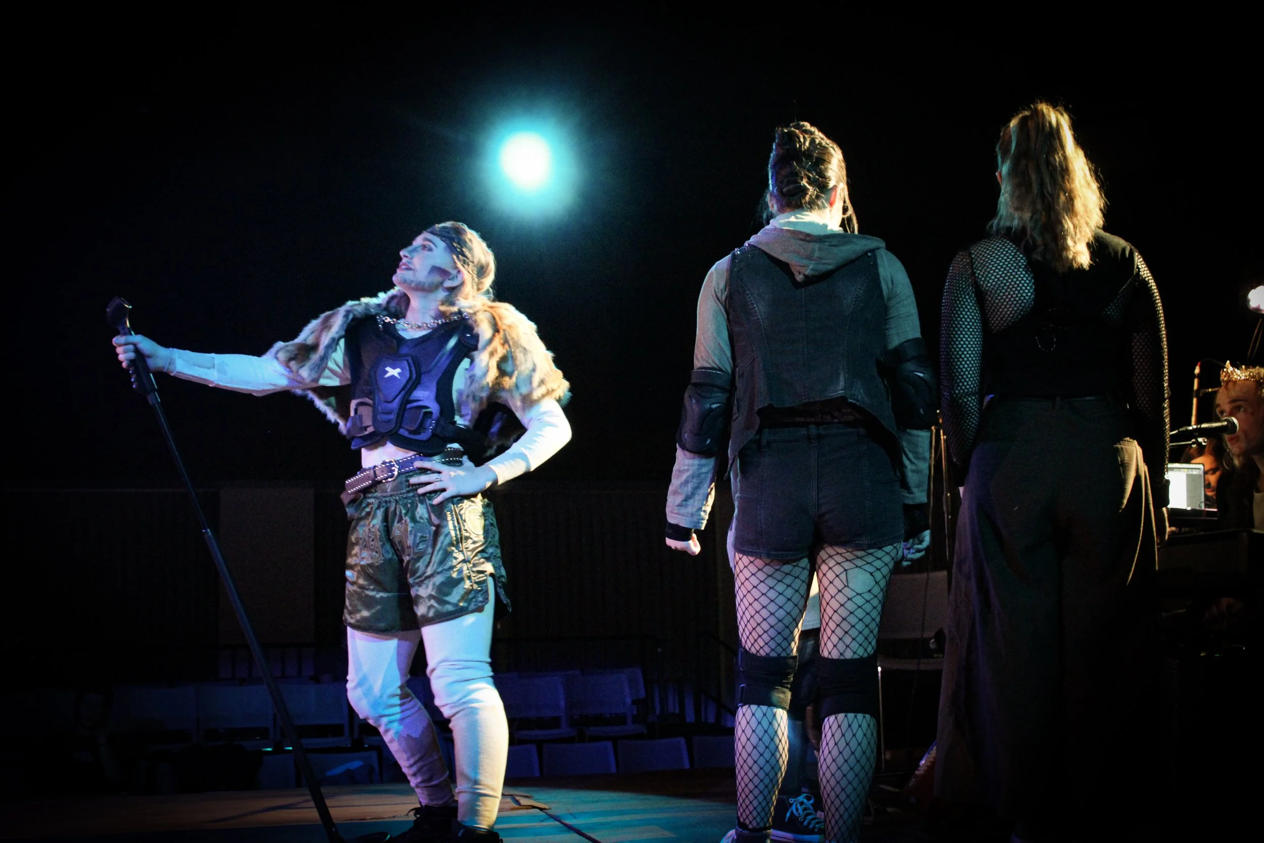 Three women on stage: one singing into a microphone, the other two facing away, with a piano and person wearing a crown in the background.
