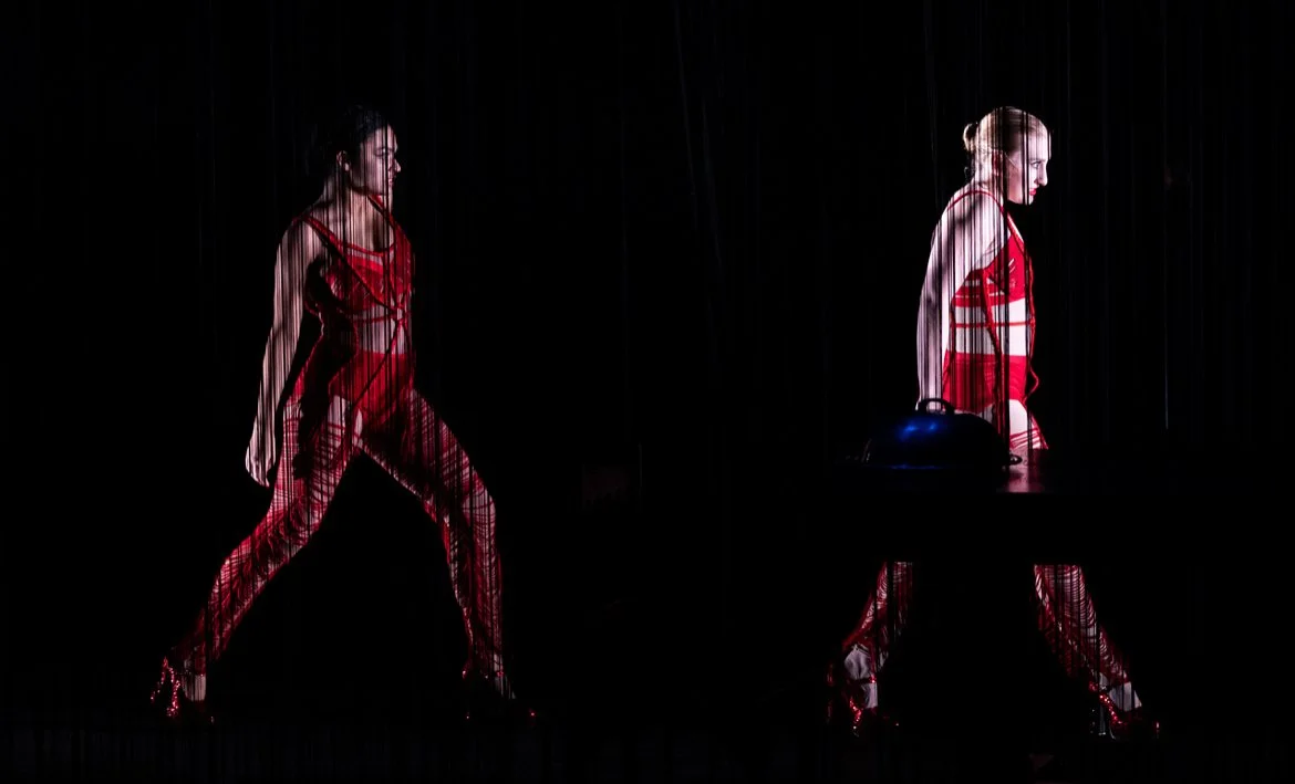 Two women dressed in red contrasting outfits, one walking and the other standing behind a desk with a black helmet or object, dimly lit against a dark background.