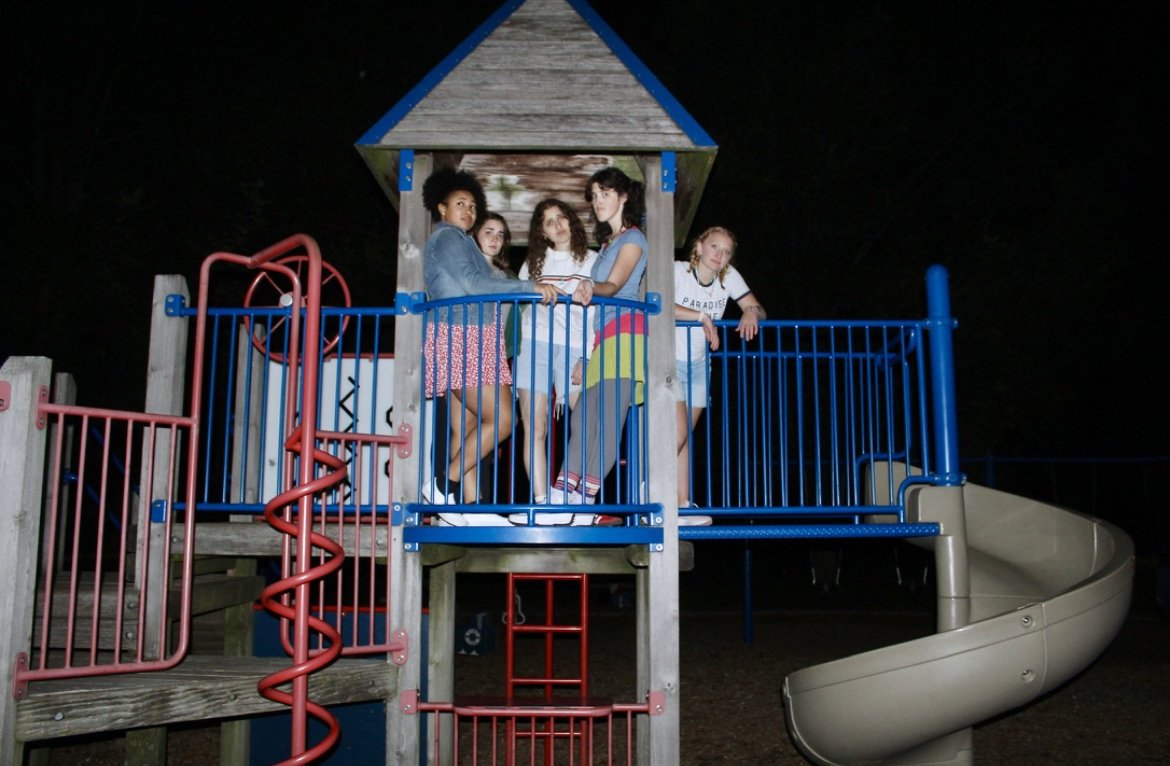 Five young girls standing on a wooden playground structure at night, with a spiral slide and blue railings.