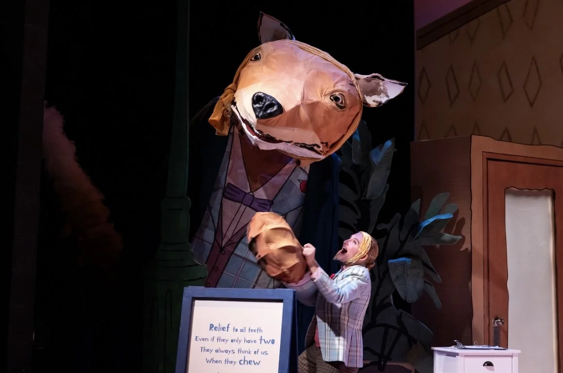 Stage scene with a person in a plaid blazer and glasses, smiling and holding a puppet of a dog's head. The puppet is oversized, with a cheerful expression, and is part of a puppet show. A sign in front reads "Relief to all teeth, Even if they only have Two, They always think of us When they chew." The background has large plants and a stylized door or wardrobe.