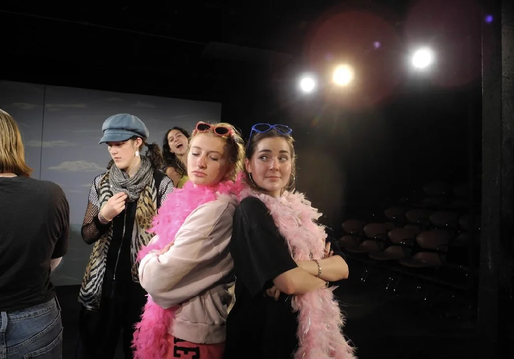 Four women at a stage or theater, two wearing pink feather boas and sunglasses, posing with crossed arms, two others in the background laughing and modeling accessories.