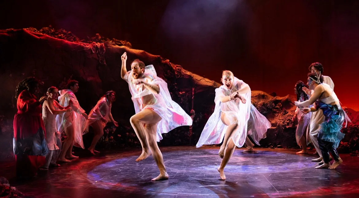 Dancers performing on stage with a rocky, volcanic backdrop, wearing flowing costumes, with two dancers jumping in the center while others watch.