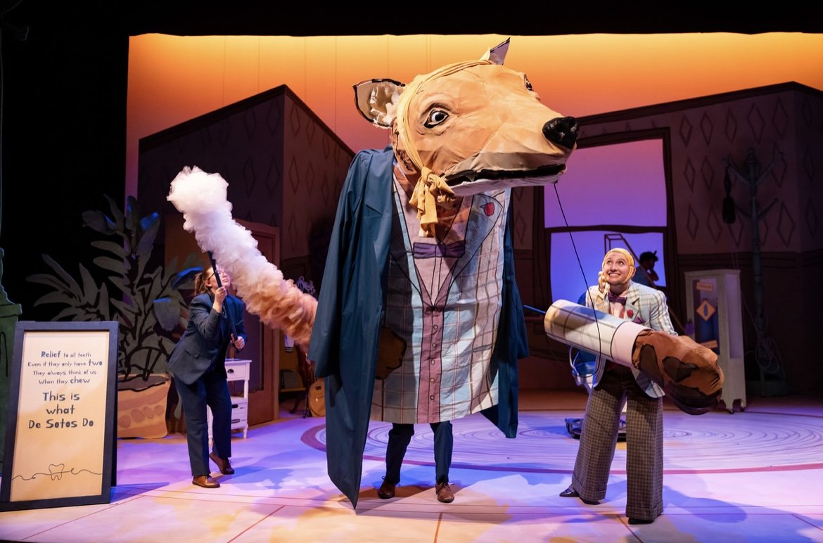 Stage scene with actors and a large dog puppet on a set designed like a house interior. The puppet has a head resembling a dog, dressed in human clothing, with a big nose and leashed tail. A sign with text about dental relief is visible on the left.