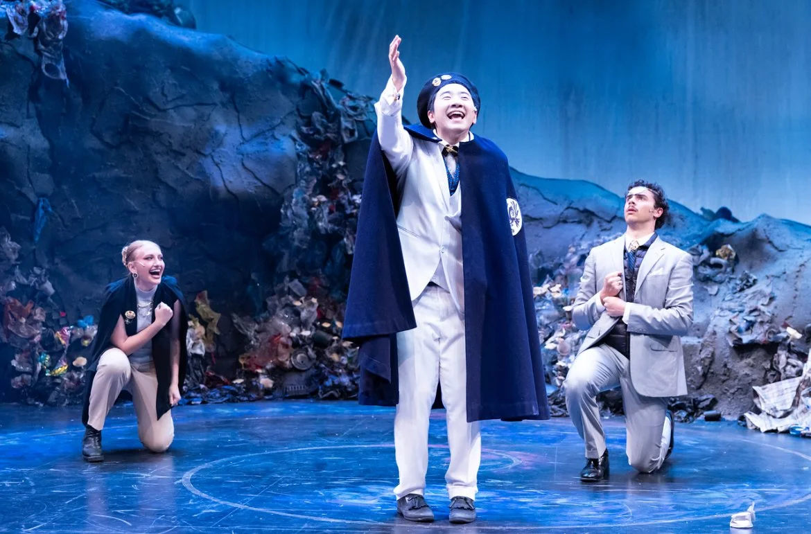 Three performers on stage during a theatrical play, with a woman on the left, a man in the center in a graduation gown, and another man on the right, set against a backdrop of garbage and a blue waterfall.