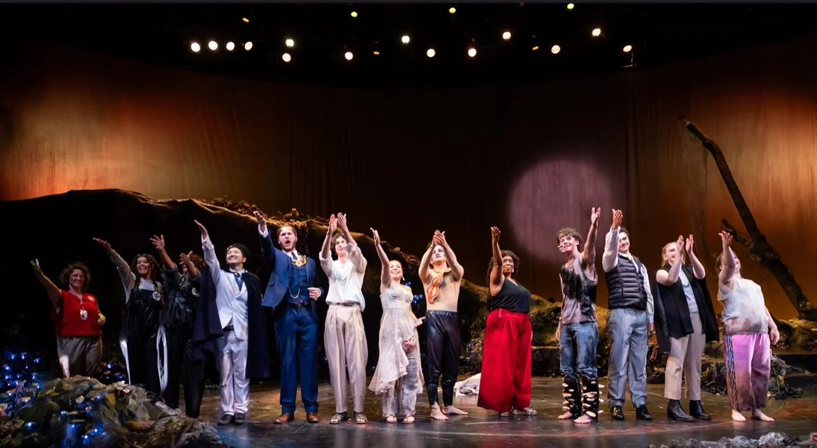Cast members on stage after a theatrical performance, holding hands and taking a bow, with stage scenery and lighting in the background.