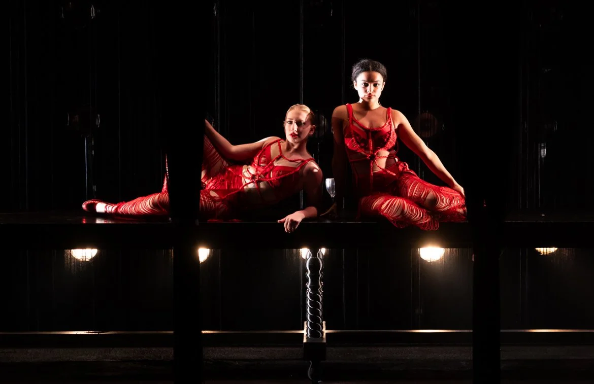 Two women in red costumes sitting on the floor in front of a dark background, illuminated by stage lights.