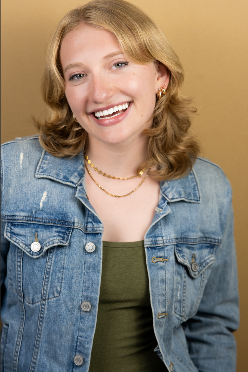 Smiling young woman with blonde hair, wearing a denim jacket, green top, and gold jewelry, posed against a beige background.