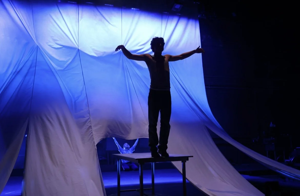 Performer balancing on a table with arms outstretched, backlit by blue lighting, with a person in the background with raised hands and fabric draping overhead.