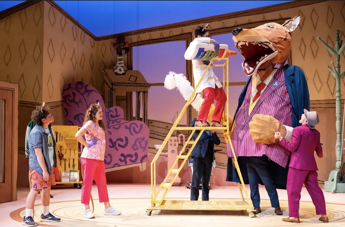 Children and adults in costume rehearsing a scene from a play with a giant anthropomorphic dog wearing a suit, with a child in a lion mask and costumes and others. The scene is set in a colorful, cartoon-like indoor stage.