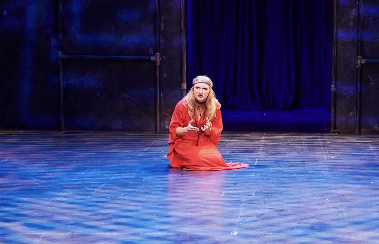 A woman in a red dress and headband kneeling on stage with a dark backdrop.