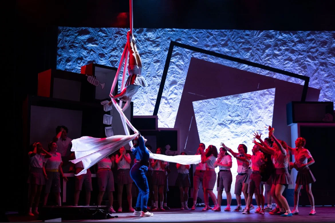 Stage performance with acrobatic aerial silk act and group of performers dressed in white and gray standing and reaching out, with abstract geometric backdrop.