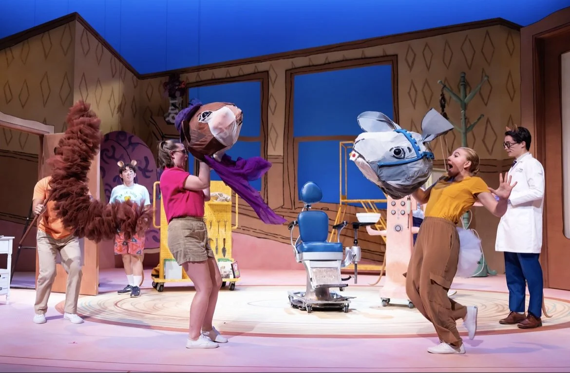 Children on stage wearing animal head costumes, including a bear, a fish, and a rabbit, as part of a playful theater production set resembling a veterinarian's office.
