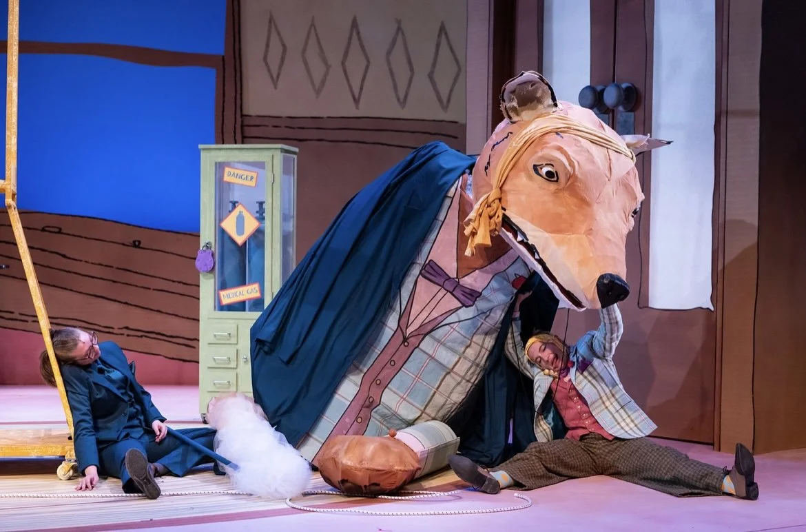 Two children in school costumes interacting with a large puppet of a dog with a yellow scarf, set on a colorful stage with a backdrop resembling a wooden room.