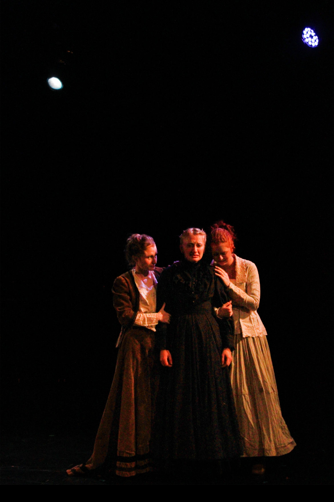 Three women in period costumes performing on a dark stage with stage lights above.