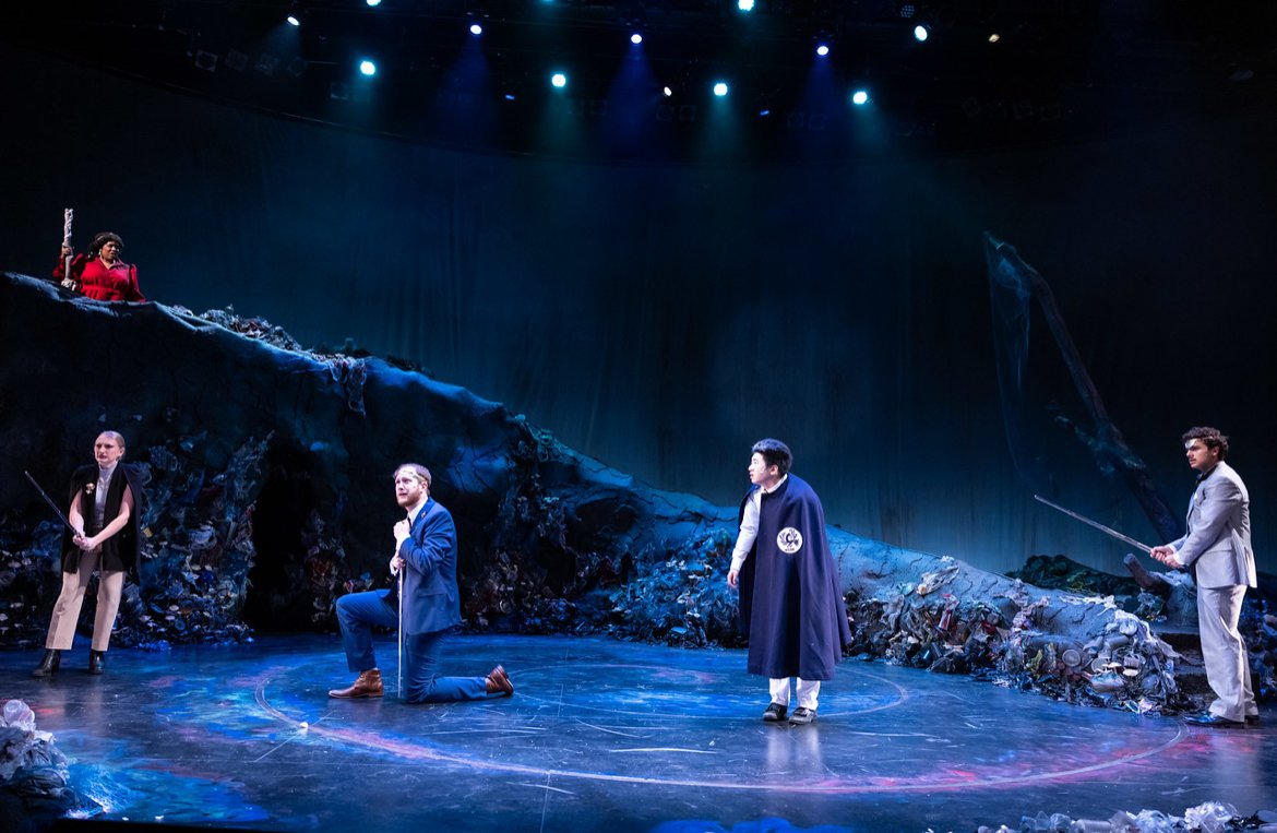 Theatrical performance on stage depicting a scene with five actors, some kneeling and others standing, against a backdrop resembling a pile of trash with a dark and blue-lit setting.