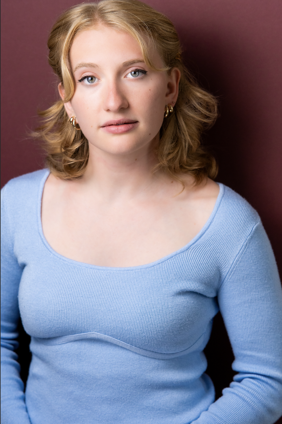 A young woman with light skin, curly blonde hair, and blue eyes wearing a light blue sweater, standing against a dark pink background.