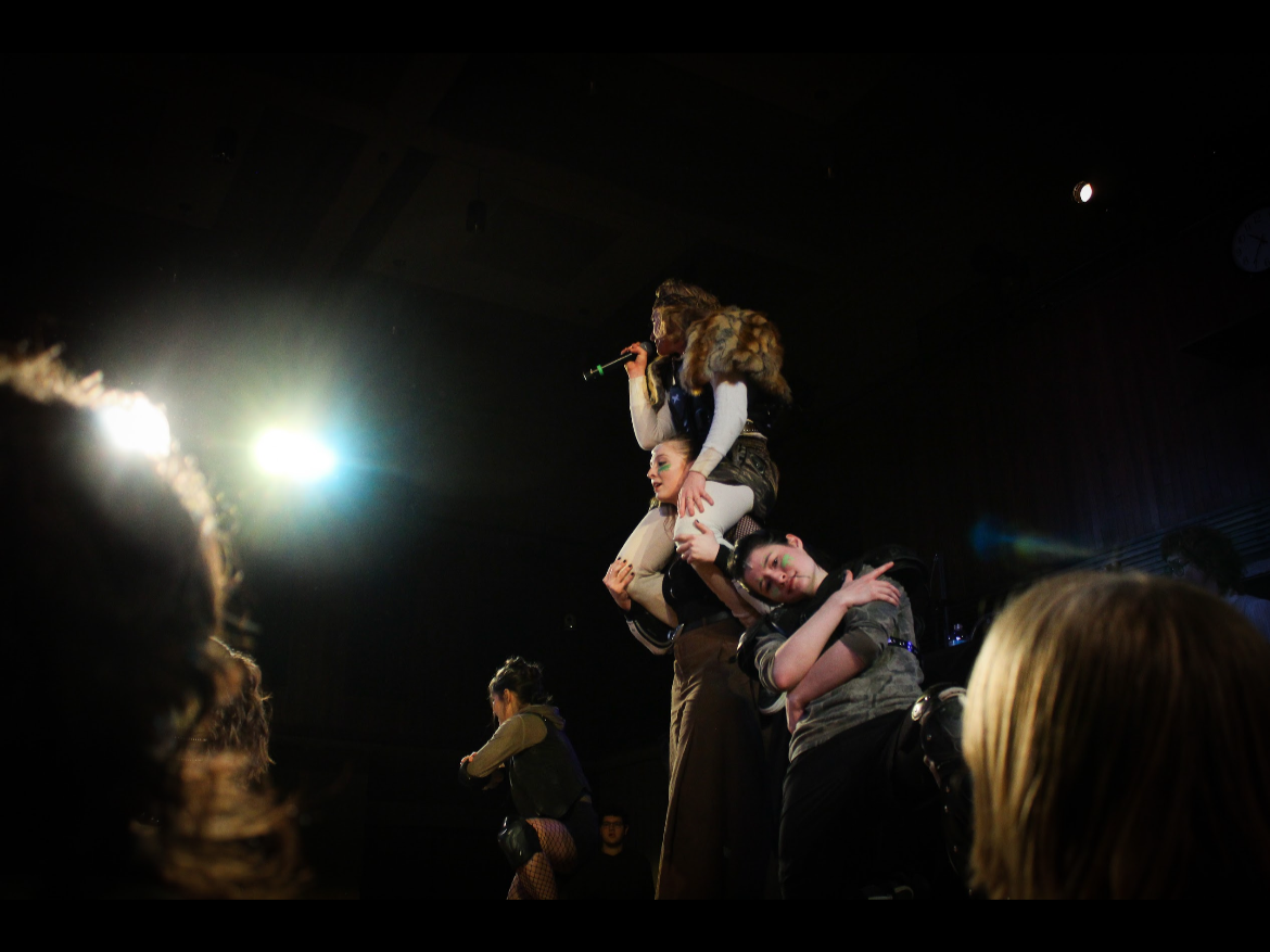 Performer stands on shoulders of another performer during a stage act, with audience members watching in the foreground and stage lighting overhead.