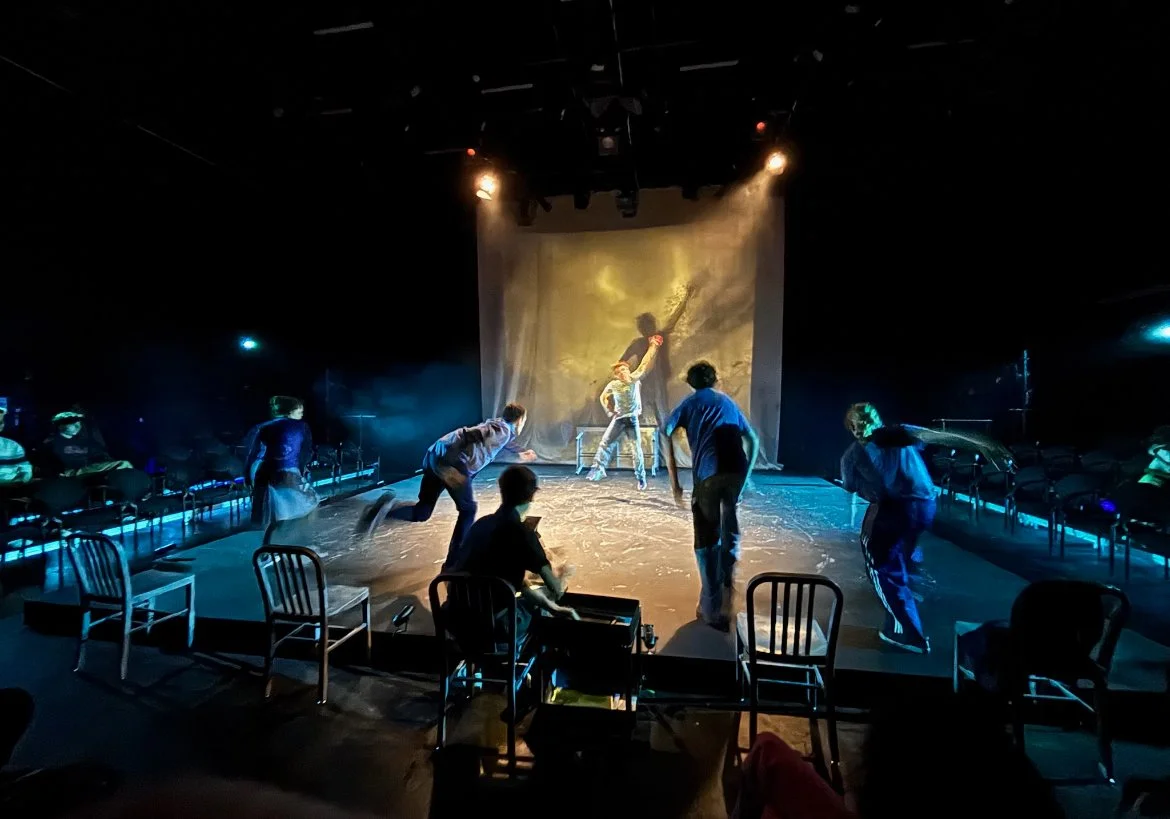 Theater stage with actors performing a scene, dim lighting, and audience seats visible in the foreground.