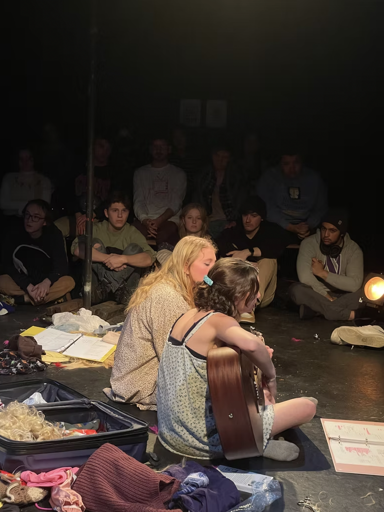 A young girl with shoulder-length brown hair sits on the floor playing a guitar, with a blonde woman beside her, surrounded by an audience of teenagers seated on the floor, attentively watching a performance in a dark room.