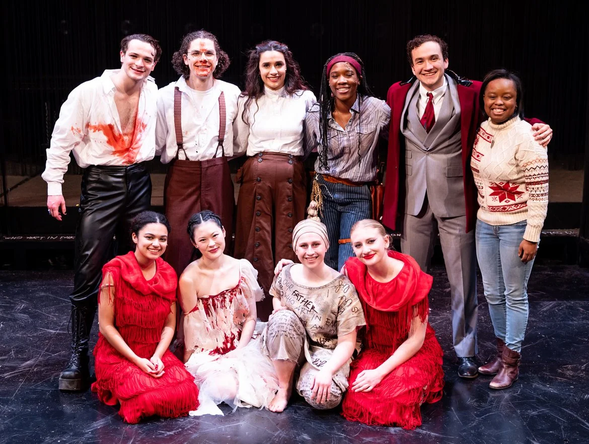 Group of ten actors on stage after a rehearsal or performance, smiling and posing for a photo, dressed in costume with some wearing makeup and accessories.