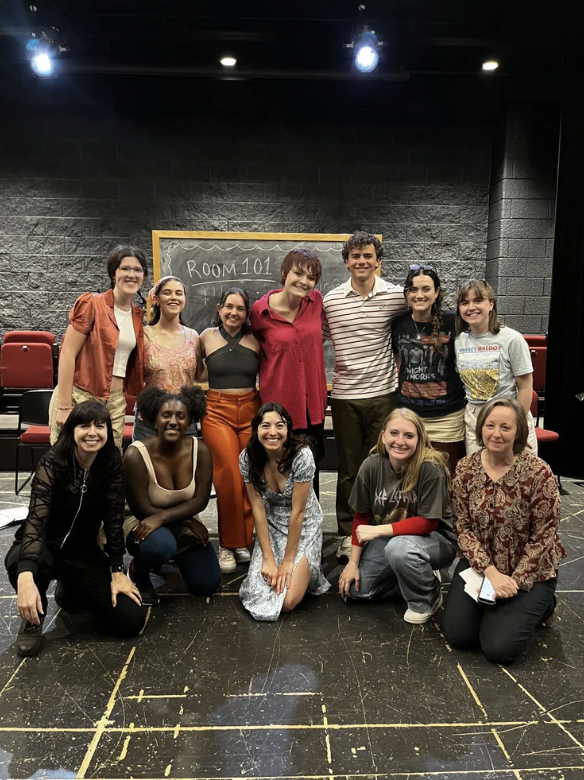 Group of eleven diverse people posing together on a theater stage, with a blackboard behind them that reads 'ROOM 101'.