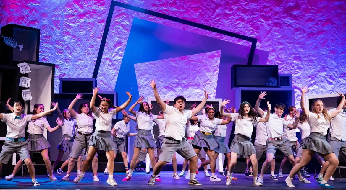 A group of children dressed in school uniforms performing on stage with colorful abstract background.
