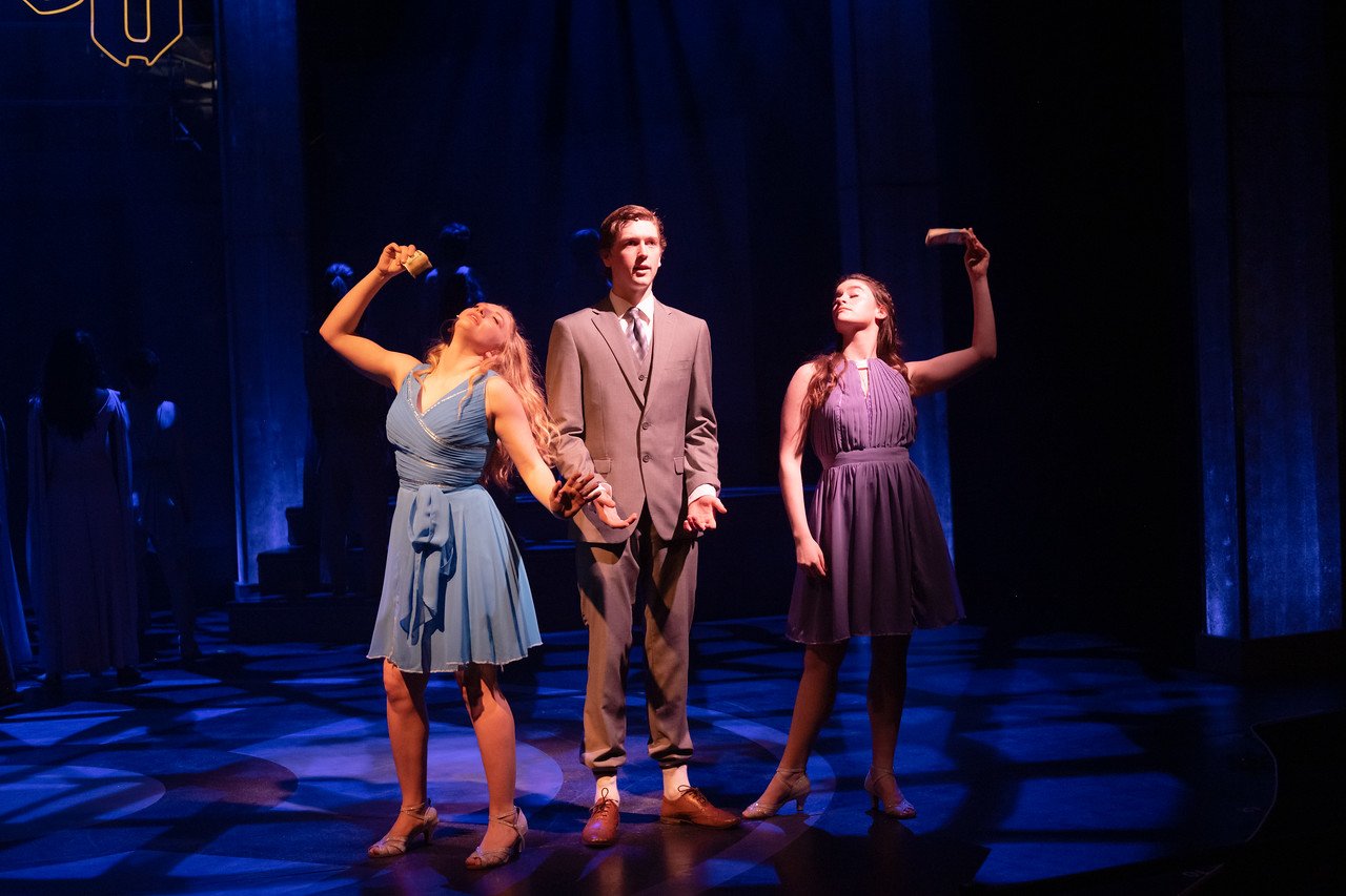 Three young actors performing on stage under dramatic lighting, with two women in dresses and a man in a suit, possibly during a theatrical production.