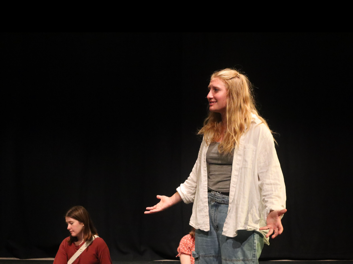 A woman with long blonde hair speaking on stage, gesturing with her hands. She is wearing a white shirt over a gray top and blue jeans. Two other women are seated nearby, one wearing a red top and the other partially visible.