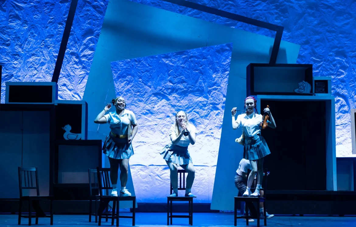 Three women performing on stage, standing on chairs, in a room with blue and white textured walls and shelves with various items.