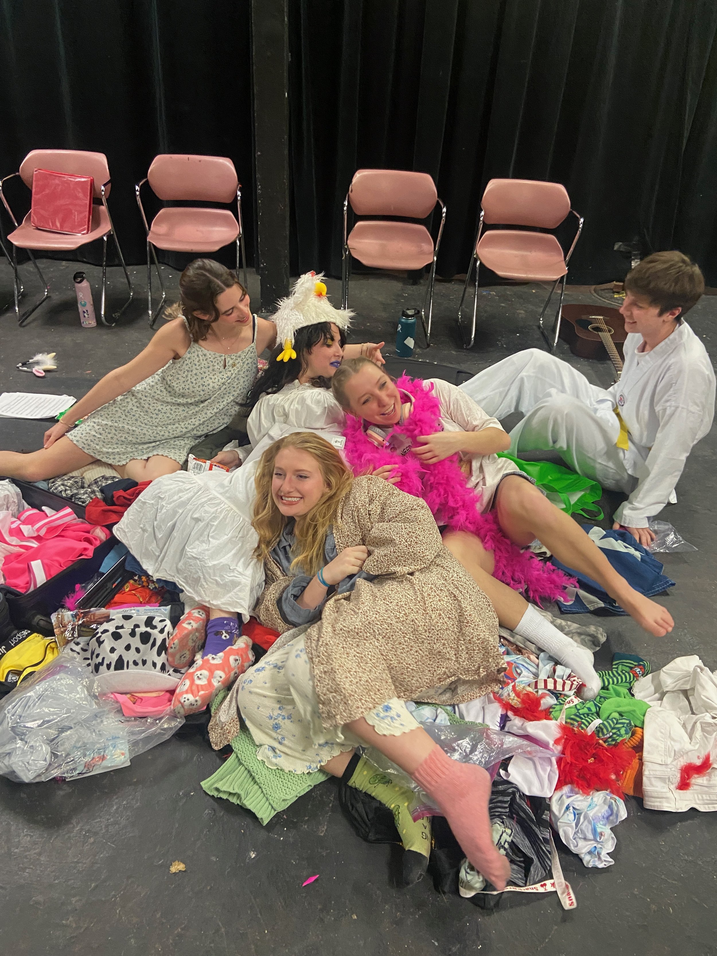 Five young women dressed in costumes and casual clothing are lying on a stage floor among clothes and props, smiling and enjoying each other's company.
