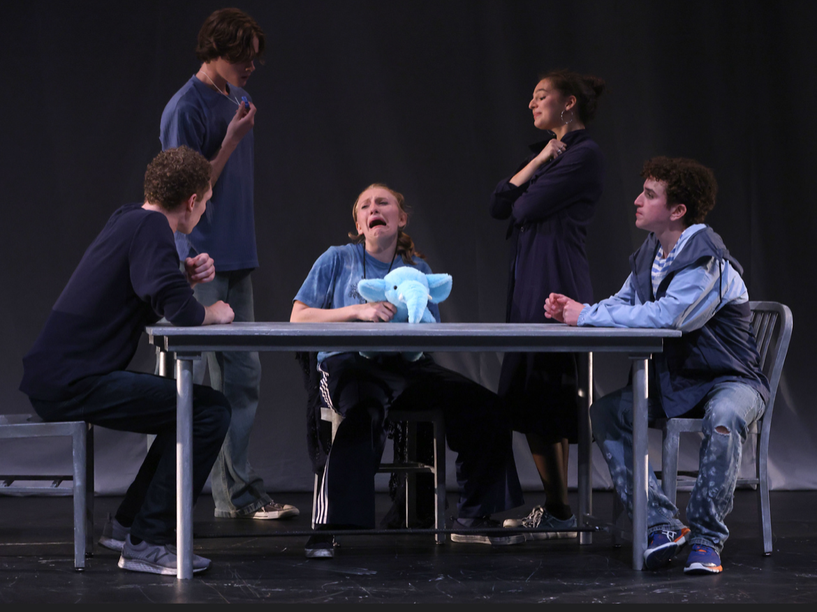 The scene depicts five young people on stage, engaged in a dramatic scene. One girl in the center is crying, holding a blue stuffed elephant, with tears on her face. Two young men are seated on either side of her, facing her and looking distressed. Two girls are standing behind the seated boys, with one looking down at her phone and the other with her arms crossed, appearing contemplative.