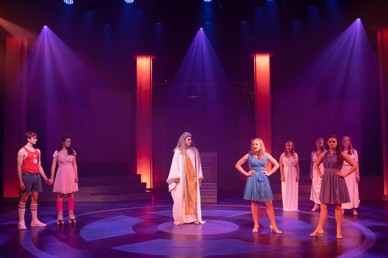 Theater stage with actors in colorful costumes, some holding hands, under purple and red stage lighting.