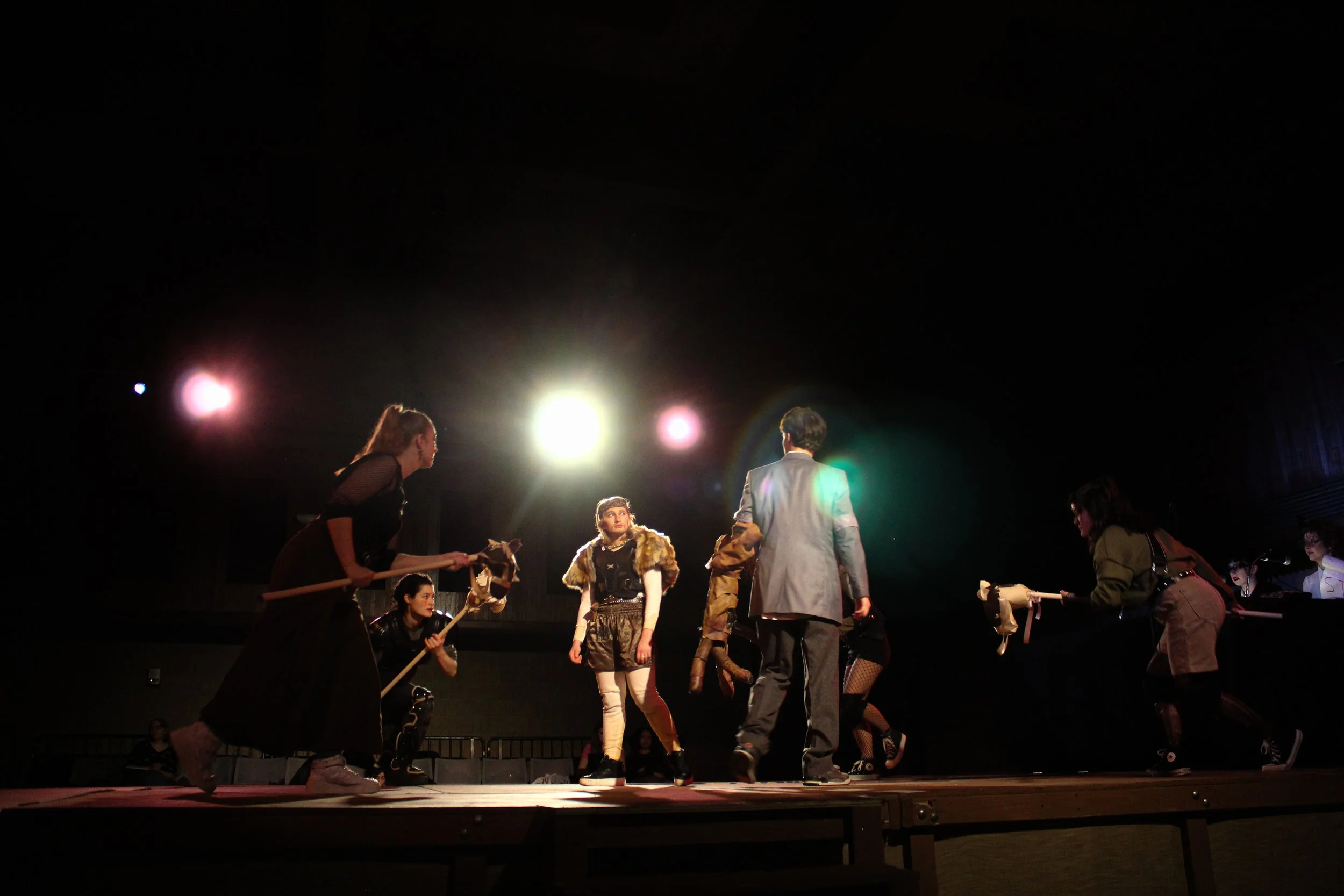 Theatrical scene with performers, some dressed in costumes and holding props, on stage under bright lights in a dark auditorium.