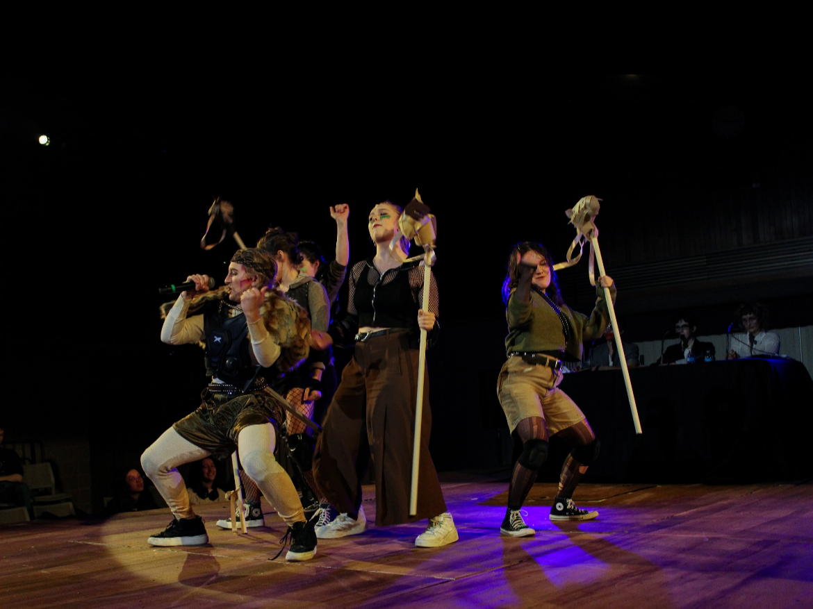 Group of people performing on stage, some holding wooden sticks with plush animal heads, in a dark theater setting.