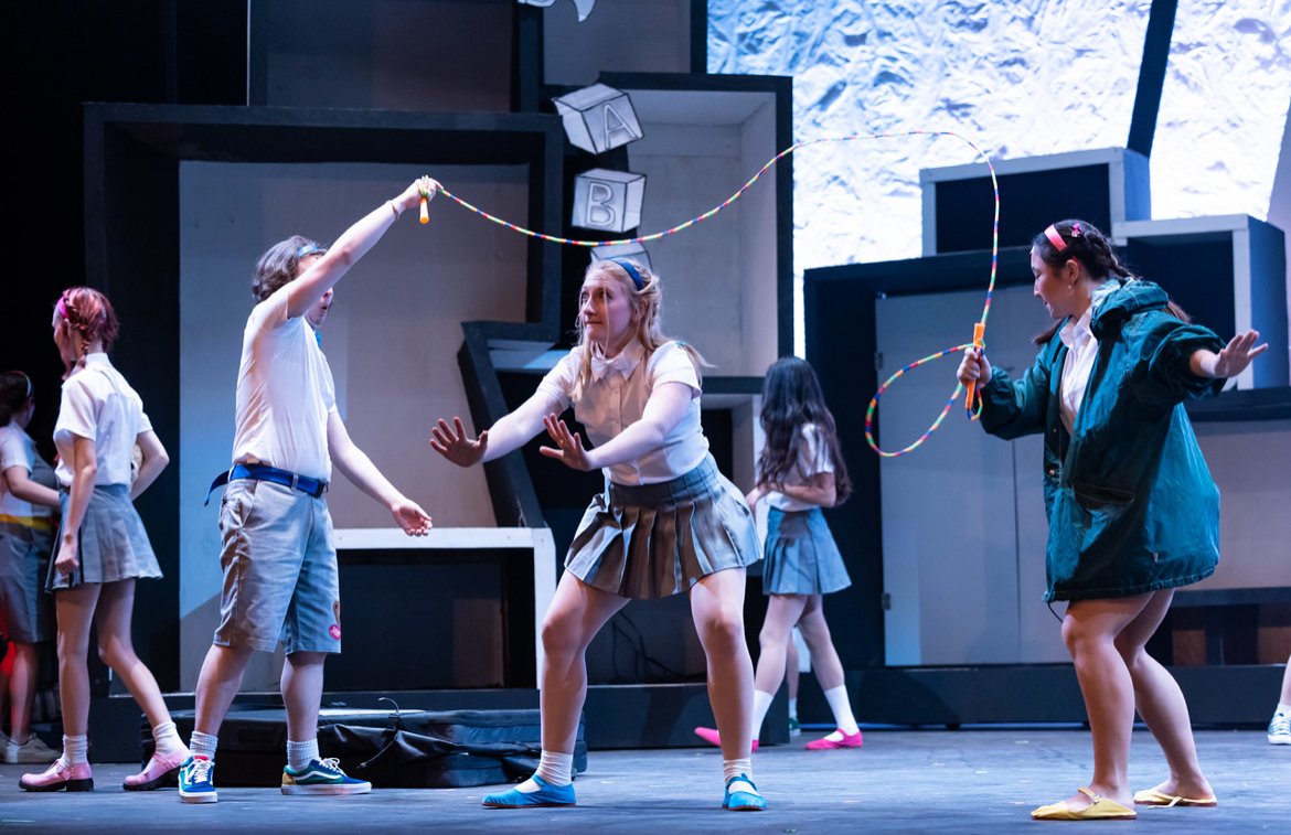 Children acting in a school play on stage, some holding jump ropes, with set pieces and blocks labeled A and B in the background.