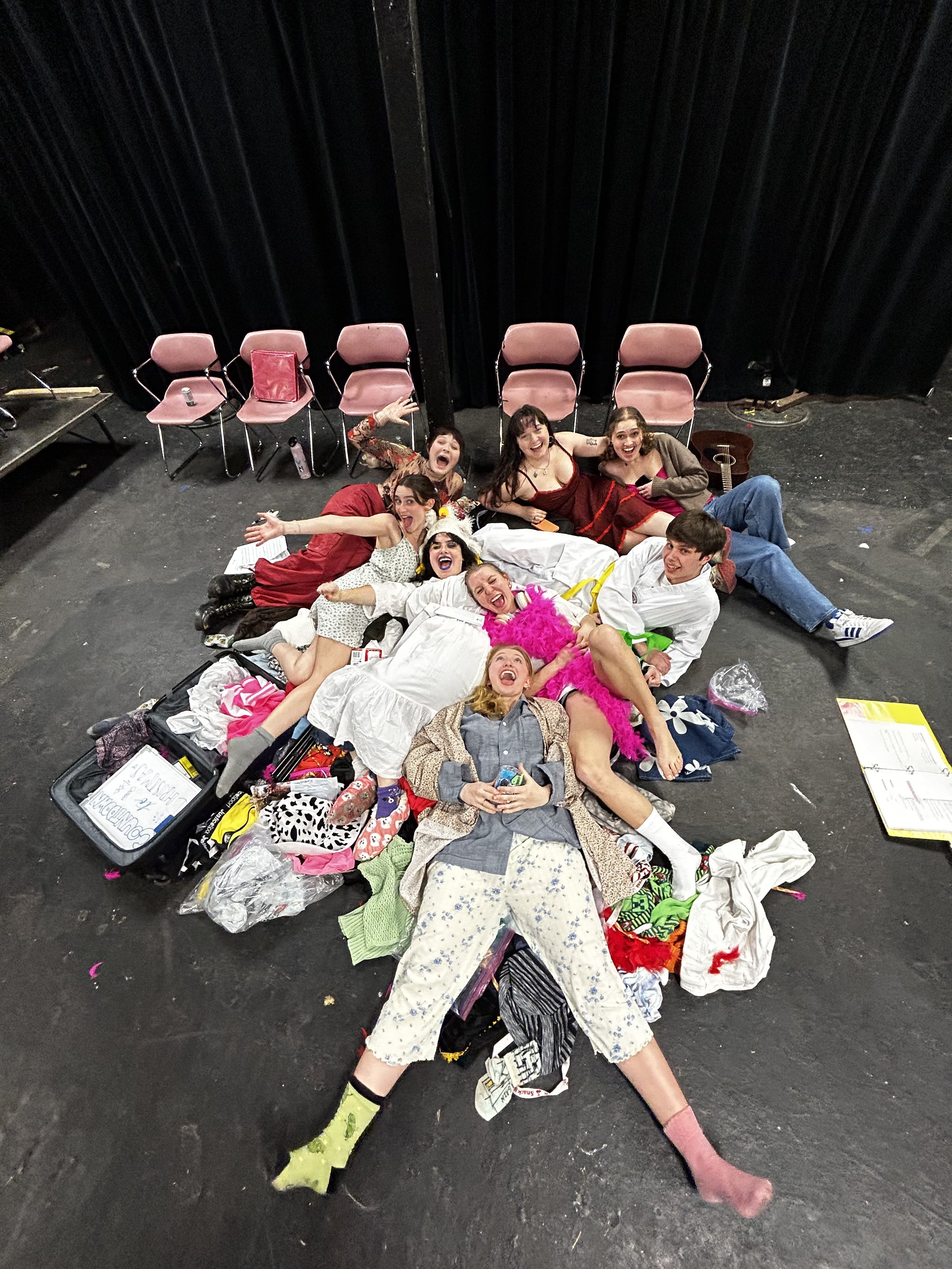 Group of people lying on stage floor surrounded by clothes and props, celebrating and smiling, with empty chairs and black curtains in the background.